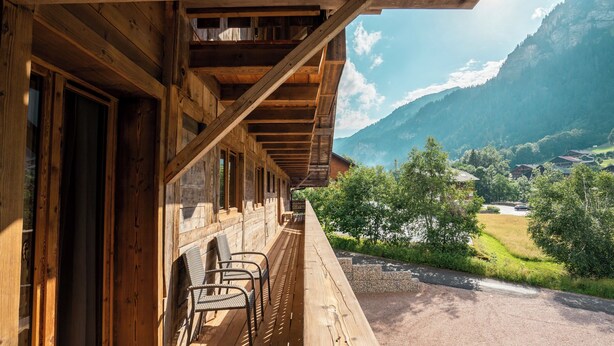 A wooden balcony with chairs, overlooking a mountainous landscape with houses and greenery.