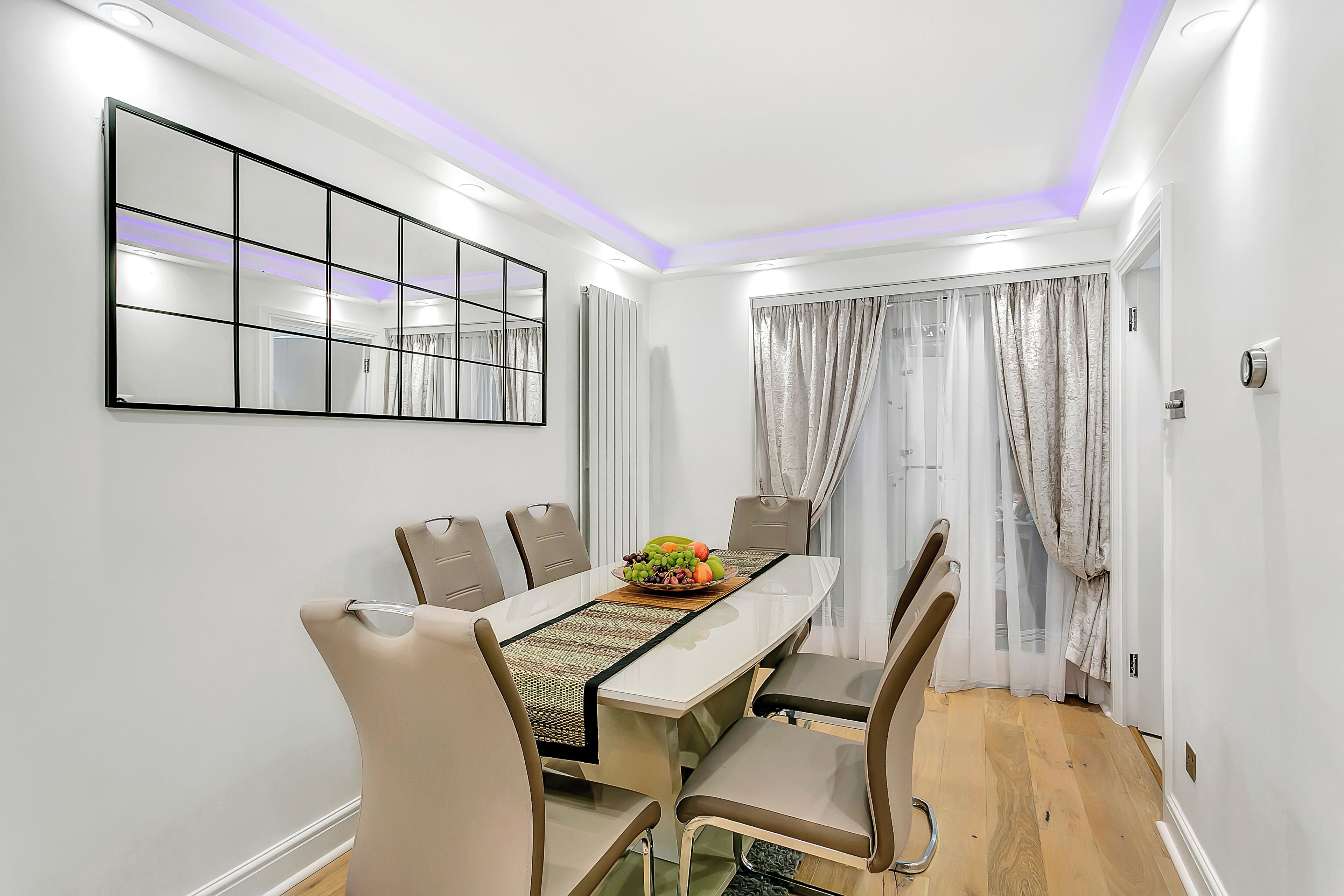 A dining area with a white table, beige chairs, a fruit bowl, and a mirror on the wall.