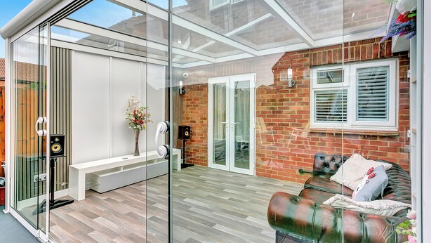 A modern patio with a glass enclosure, a cushioned seating area, and a brick wall.