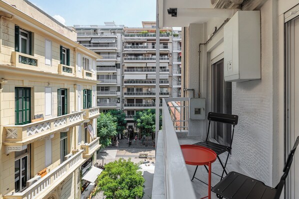 Un balcon avec une table et des chaises rouges, donnant sur une cour intérieure agrémentée d’arbres et sur un immeuble doté de nombreux balcons.