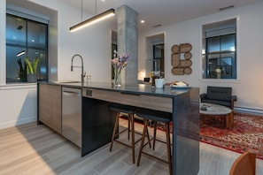 A modern kitchen with a central island, bar stools, a coffee machine, and a dining area with a sofa and coffee table.