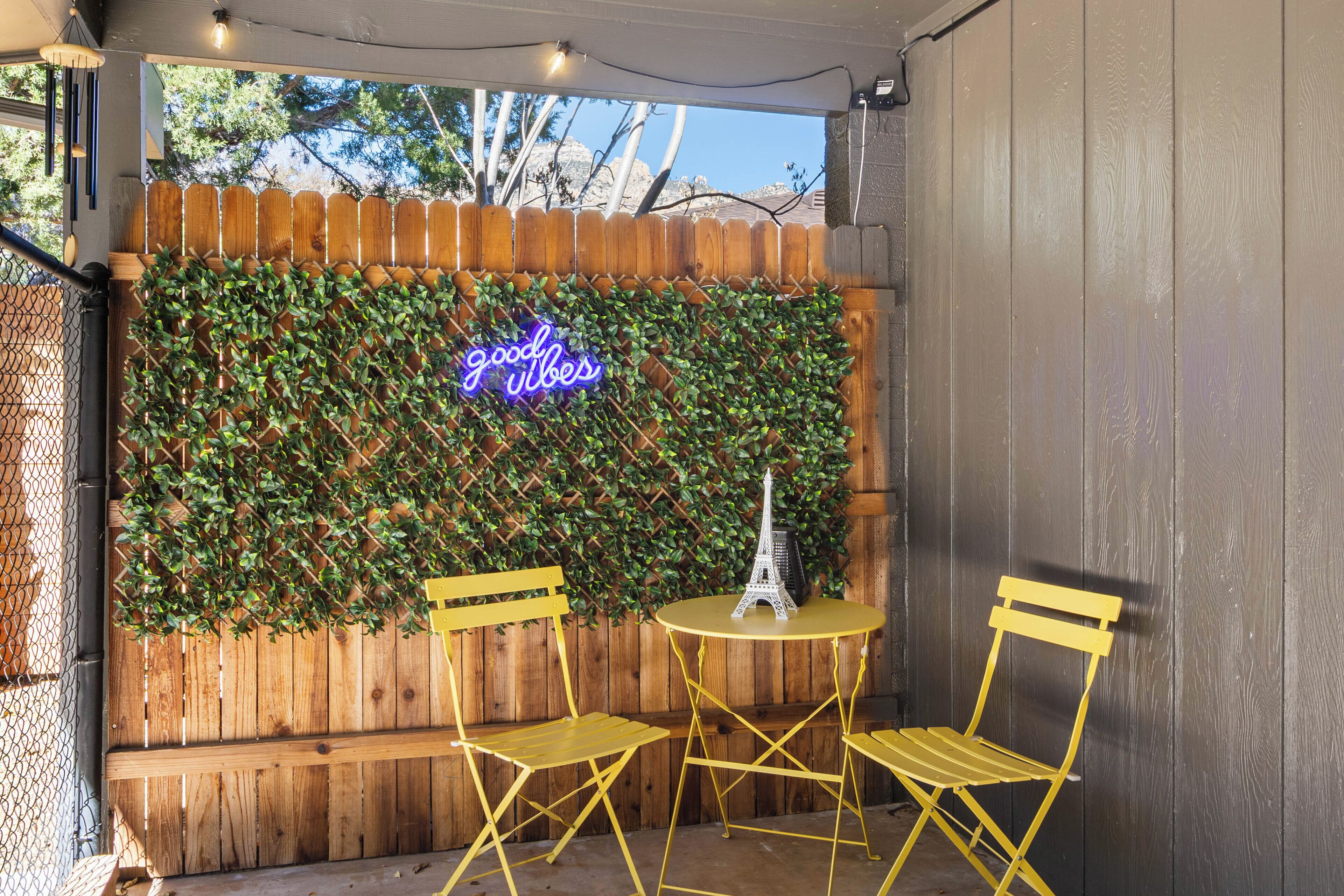 An outdoor seating area with yellow chairs and a small round table, a neon sign, and a wooden fence with greenery.