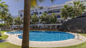 Uma piscina rodeada por palmeiras e uma construção com telhado de palha, com um edifício de vários andares ao fundo.