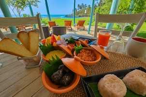 Uma área de refeições ao ar livre tropical com vista para o oceano, apresentando diversos pratos em tigelas de cerâmica e um copo de suco de laranja.