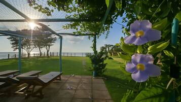 A covered outdoor seating area with picnic tables, a view of the sea, and purple flowers in the foreground.