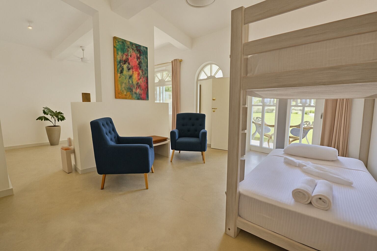 Ein Hotelzimmer mit einem Etagenbett, zwei blauen Sesseln, einem kleinen Tisch, einer Topfpflanze und einem farbenfrohen, abstrakten Gemälde an der Wand.