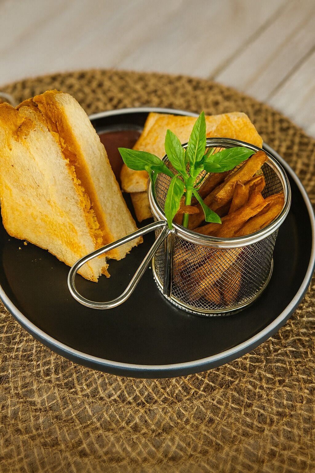 A black plate with a metal strainer containing fried items and a sprig of mint, accompanied by a slice of bread on a textured surface.