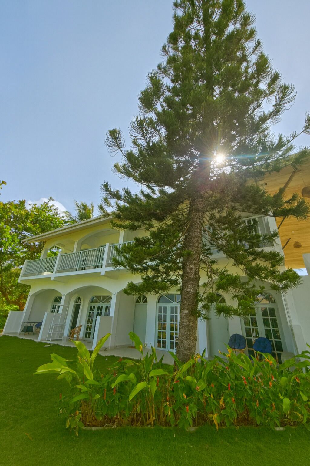 A two-story house with a balcony, surrounded by lush greenery and a large tree.