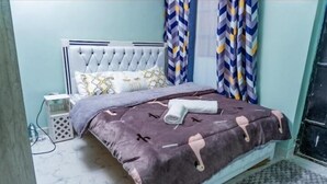 Un dormitorio con cabecera capitonada, cama con manta estampada, mesita de noche y cortinas.
