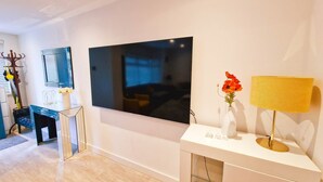 A modern living room with a large flat-screen TV mounted on the wall, a white console table with a vase of flowers, and a yellow lamp.