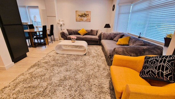 A modern living room with a grey sofa, yellow armchair, and a white coffee table. There is a dining area with a table and chairs, and a kitchen area with a fridge and cabinets.