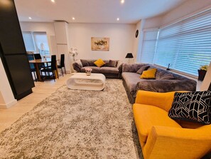 Ein modernes Wohnzimmer mit grauem Sofa, gelbem Sessel und weißem Couchtisch. Es gibt einen Essbereich mit Tisch und Stühlen sowie eine Küche mit Kühlschrank und Schränken.