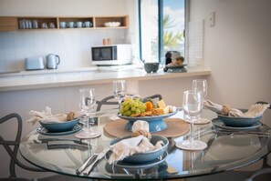 A modern kitchen with a dining table set for a meal, featuring a bowl of fruit, wine glasses, and neatly folded napkins. - Blue Beyond Airlie (Airlie Beach)
