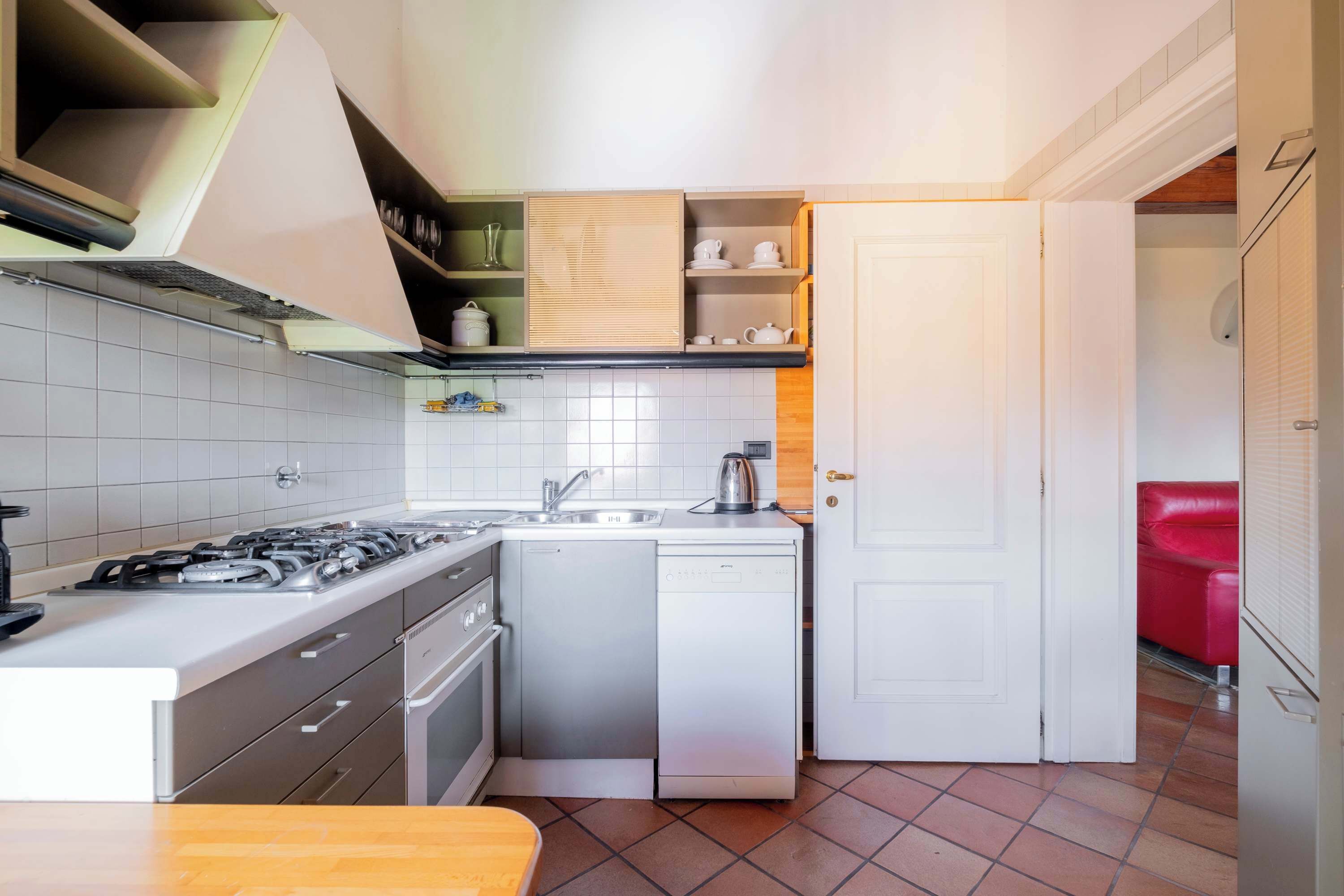 Cocina compacta con estufa a gas, lavavajillas y un lavamanos pequeño. Hay una mesa de madera y una silla roja en la esquina.