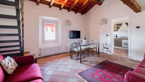 A spacious living area with a high ceiling, a dining table with chairs, a TV mounted on the wall, and a view into the kitchen.