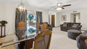 A modern living room with a glass dining table, two sofas, a ceiling fan, and a large window with curtains. - 55+ Comfort & Quiet | 2BR Peaceful Home (Sun City West)