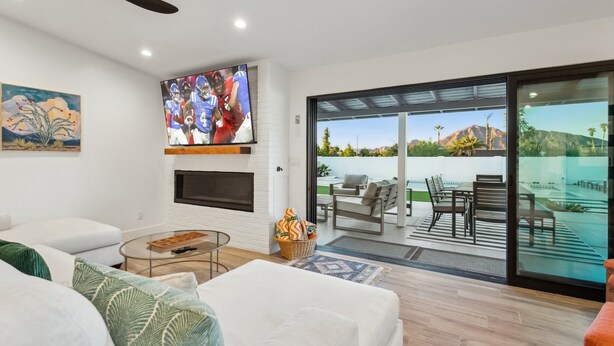 Una sala moderna con televisor de pantalla plana, chimenea, mesa de centro de vidrio y vista a un patio con mobiliario exterior.