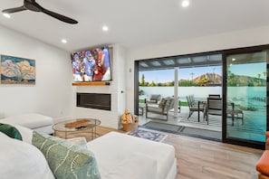 Una sala moderna con televisor de pantalla plana, chimenea, mesa de centro de vidrio y vista a un patio con mobiliario exterior.
