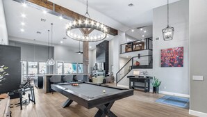 A modern living room with a pool table, a dining area, and a staircase leading to an upper level.
