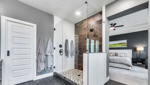 A modern bathroom with a glass shower enclosure, a white door, and a hallway leading to a bedroom with a bed and a nightstand.
