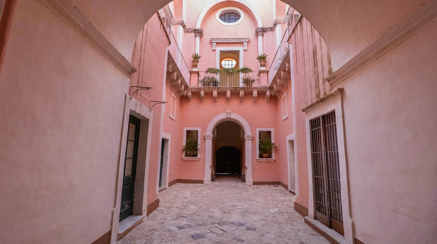 Ein schmales, in Rosa gehaltenes Innenhof mit einem zentralen Bogen, der in einen dunklen Innenraum führt.