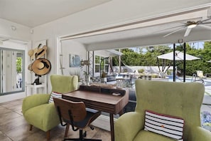 Una habitación con una mesa de comedor, dos sillones verdes, una silla de madera, un ventilador de techo y vistas a una piscina.