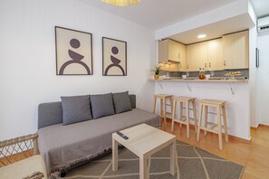A modern living room with a grey sofa, a small wooden table, and a kitchen area with wooden cabinets and stools.