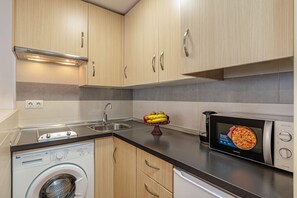 A modern kitchen with wooden cabinets, a washing machine, a microwave, and a fruit bowl on the counter.