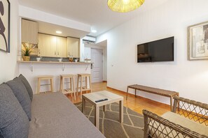A modern living room with a sofa, a coffee table, a TV, and a kitchen area with wooden cabinets and a hanging plant. - Bermeja Village | Balcony & Scenic Street (Marbella)