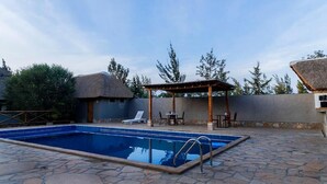 A swimming pool with lounge chairs and a pergola, surrounded by stone tiles and trees.