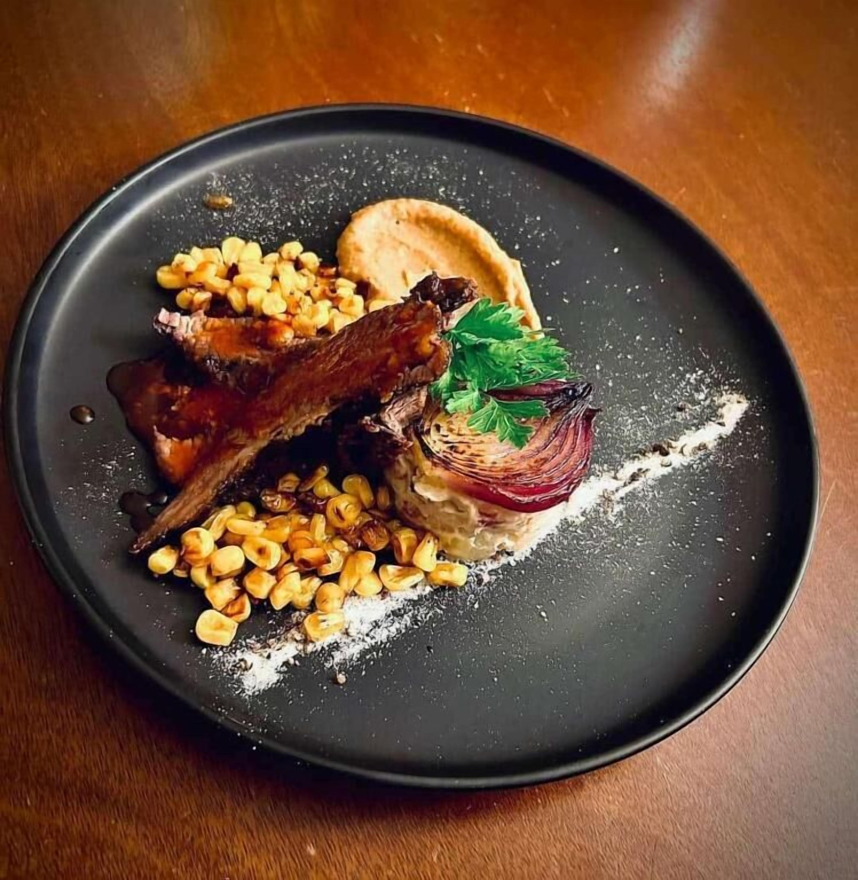 A plate with a dish consisting of meat, corn, and a garnish on a dark plate.