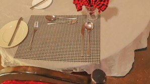 Una mesa de comedor dispuesta con platos, cubiertos y condimentos, con servilleta de cuadros rojos y negros y un pequeño jarrón con una flor roja.