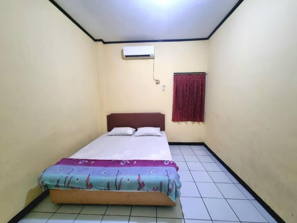 A small, single-bed room with a tiled floor, a bed with a floral bedspread, a red curtain, and an air conditioning unit on the wall.