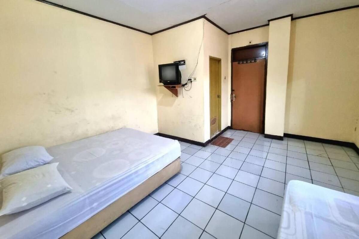 A small, simple room with a bed, a television mounted on the wall, and a door leading to another room.