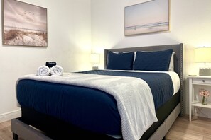 A neatly made bed with a blue and white bedspread, two pillows, and two rolled towels on the side.