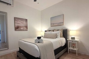 Ein ordentlich bezogenes Bett mit weißer Decke und Kissen, ein Nachttisch mit Lampe sowie ein gerahmtes Bild an der Wand.