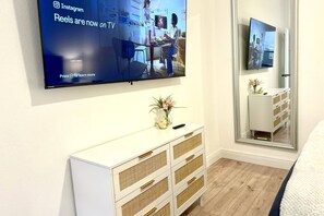 Une chambre moderne dotée d’un téléviseur à écran plat fixé au mur, d’une console blanche avec des tiroirs, d’un miroir et d’un plancher en bois.