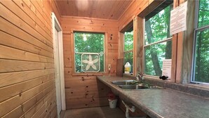 Una pequeña cocina tipo cabaña con paredes de madera, un ventilador en la ventana y un lavamanos.