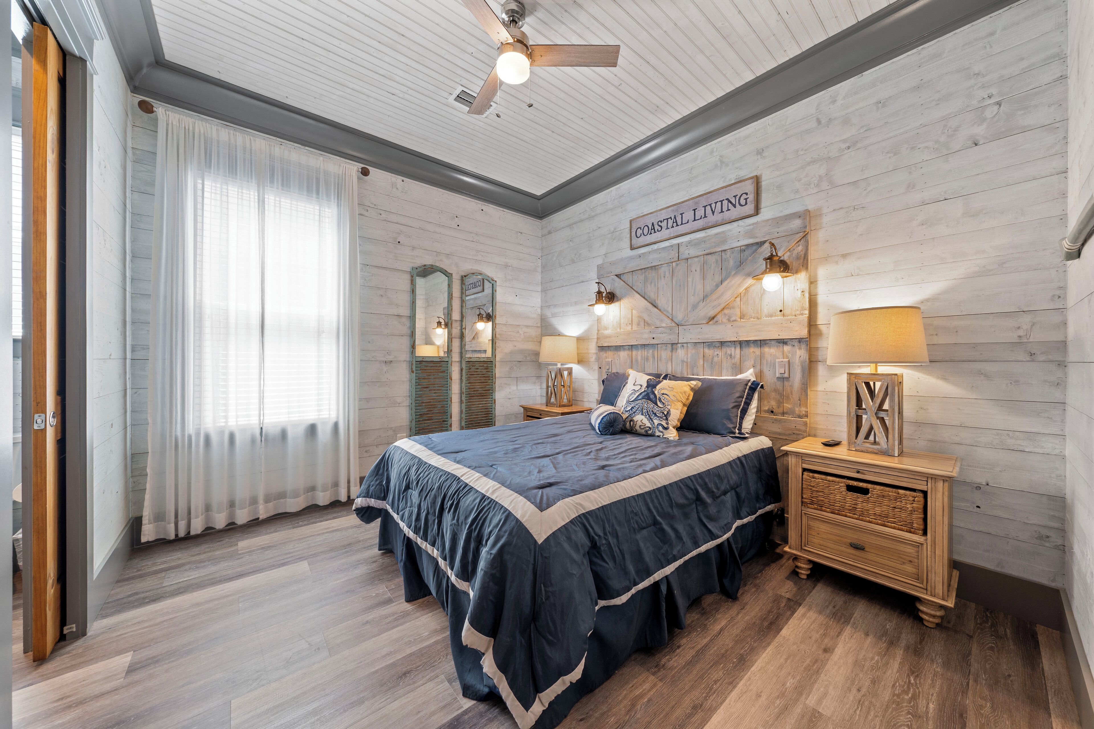 Un dormitorio con una cama grande, cabecera de madera, dos mesitas de noche con lámparas, un ventilador de techo y una ventana con cortinas de encaje.
