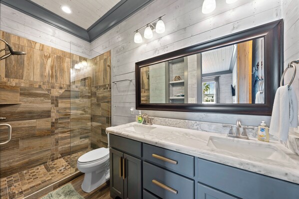 A modern bathroom with a large mirror, marble countertop, and a walk-in shower. - Southern Exposure (Orange Beach)