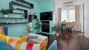 Una sala de estar moderna con paredes verde azulado, un televisor de pantalla plana, un comedor con mesa y sillas, y varias plantas en macetas.