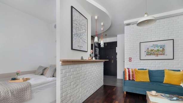 Una sala moderna con una pared de ladrillo blanco decorativa, una mesa de centro de madera, un sofá azul verdoso con cojines amarillos y una cama blanca con colcha.