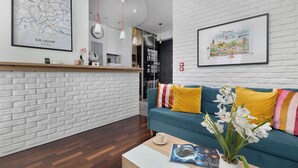 Una sala de estar moderna con una pared de ladrillo blanco, un sofá azul con cojines a rayas amarillos y rosados, una mesa de centro con un libro y un jarrón de flores blancas, y un mapa enmarcado de Cracovia en la pared.
