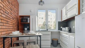 A compact kitchen with white cabinets, a brick accent wall, a window with a view of buildings, and a small trash bin.