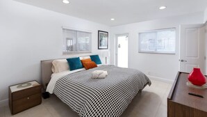 Une chambre à coucher comprenant un lit, une table de chevet, une commode et deux fenêtres équipées de stores.