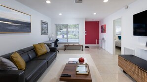 Un salon moderne avec un canapé en cuir noir, une table basse en bois et une pomme rouge posée dessus. Un téléviseur est fixé au mur et il y a une fenêtre avec des stores.