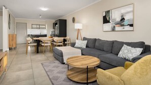 A modern living room with a grey sofa, wooden coffee table, and yellow armchair. A dining area with a wooden table and chairs is visible in the background.