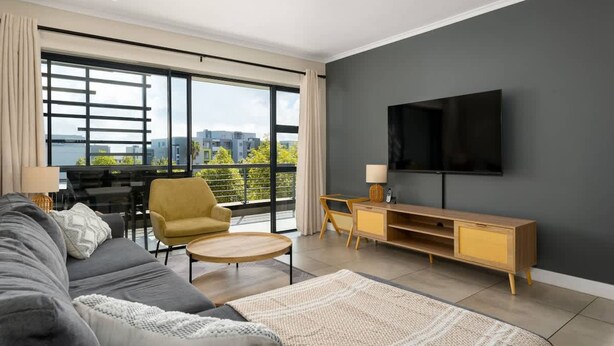 A modern living room with a sofa, armchair, and coffee table, a TV mounted on the wall, and large windows offering a city view.