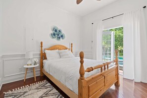 A bedroom with a wooden bed, a small bedside table with a lamp, a decorative wall piece, and a view of a pool through a glass door. - Aqua Dream - Home for 11 (St. Augustine)