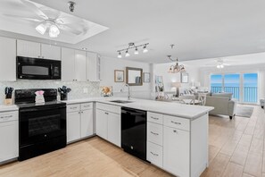 Une cuisine moderne avec des meubles blancs, un micro-ondes noir et un lave-vaisselle. On y trouve un coin repas avec une table et des chaises blanches, et une vue sur l’océan grâce à de grandes fenêtres.